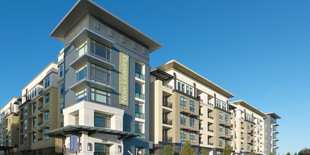 Modern multi-story apartment building with a 'Now Leasing' sign, large windows, balconies, and a clear blue sky.