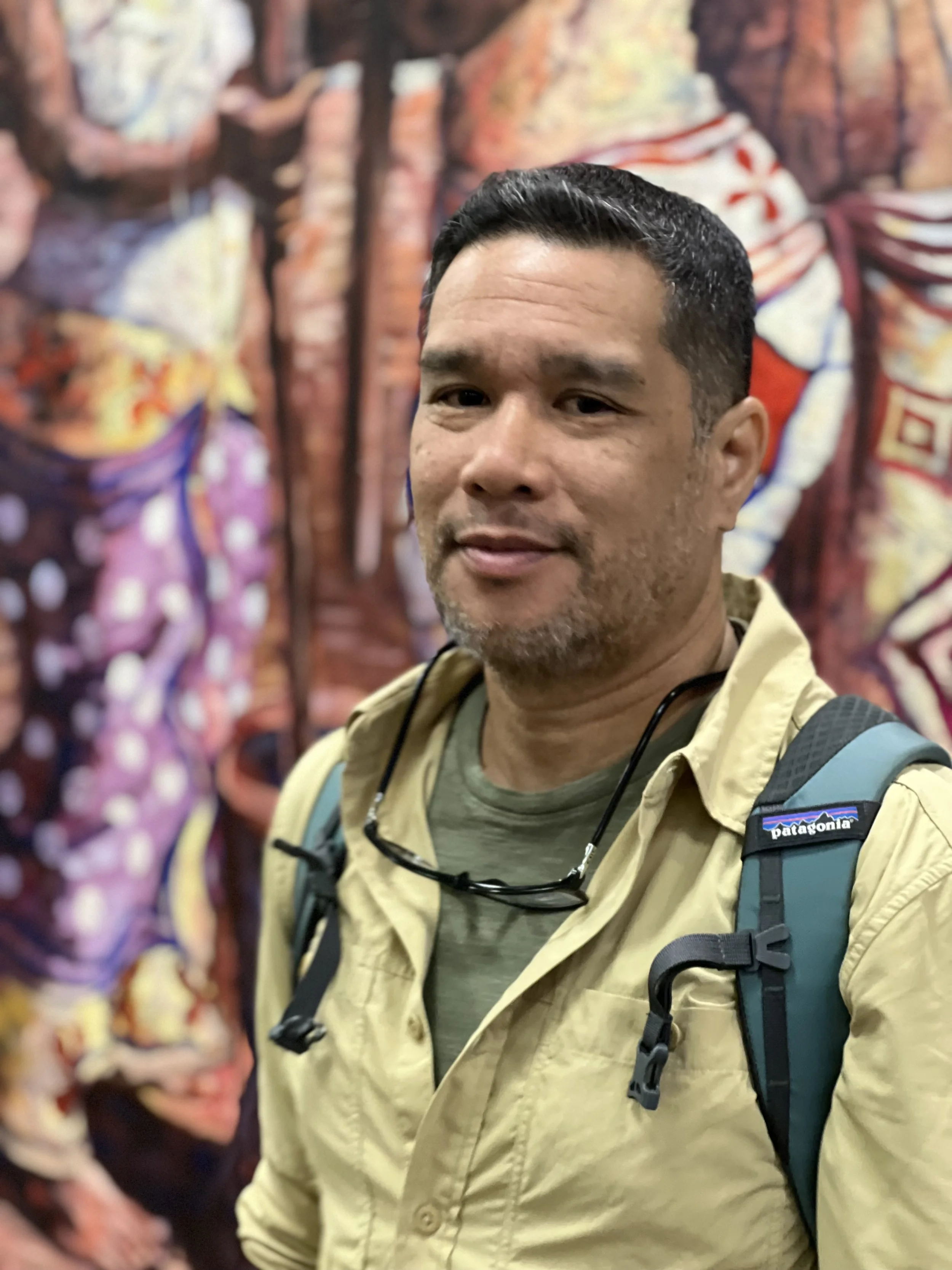A man with short black hair, wearing a tan jacket, green T-shirt, and a teal backpack, stands in front of a colorful, abstract mural.