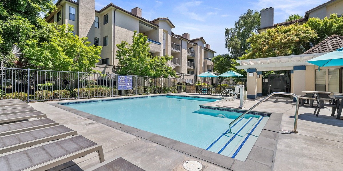 An outdoor swimming pool area in a residential complex with lounge chairs, umbrellas, a fenced perimeter, and apartment buildings in the background.