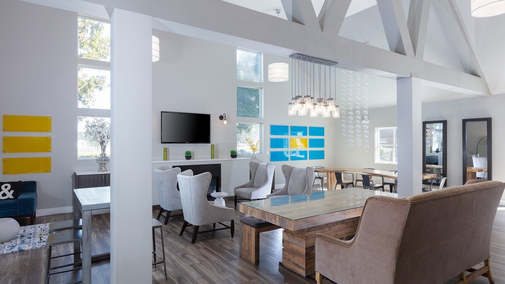 Modern living room with white walls, large windows, a flat-screen TV, and various chairs and tables, decorated with yellow and blue accents, and pendant lights hanging from the ceiling.