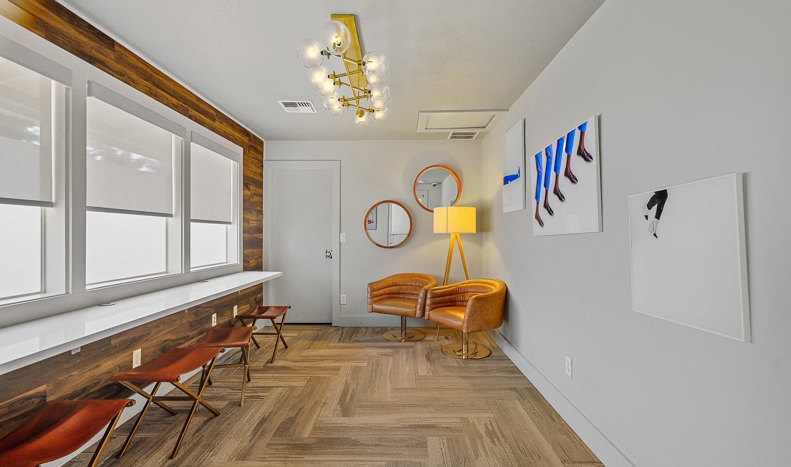 A modern interior room with wood flooring, white walls, several windows with white blinds, a gold floor lamp, two orange chairs, three round mirrors on the wall, and artwork featuring legs in blue and black clothing.