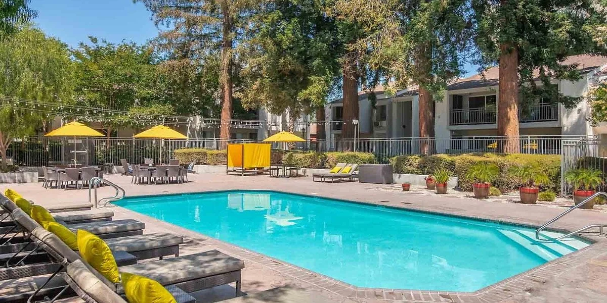 Outdoor swimming pool with lounge chairs and yellow umbrellas, surrounded by trees and greenery, in a residential complex.
