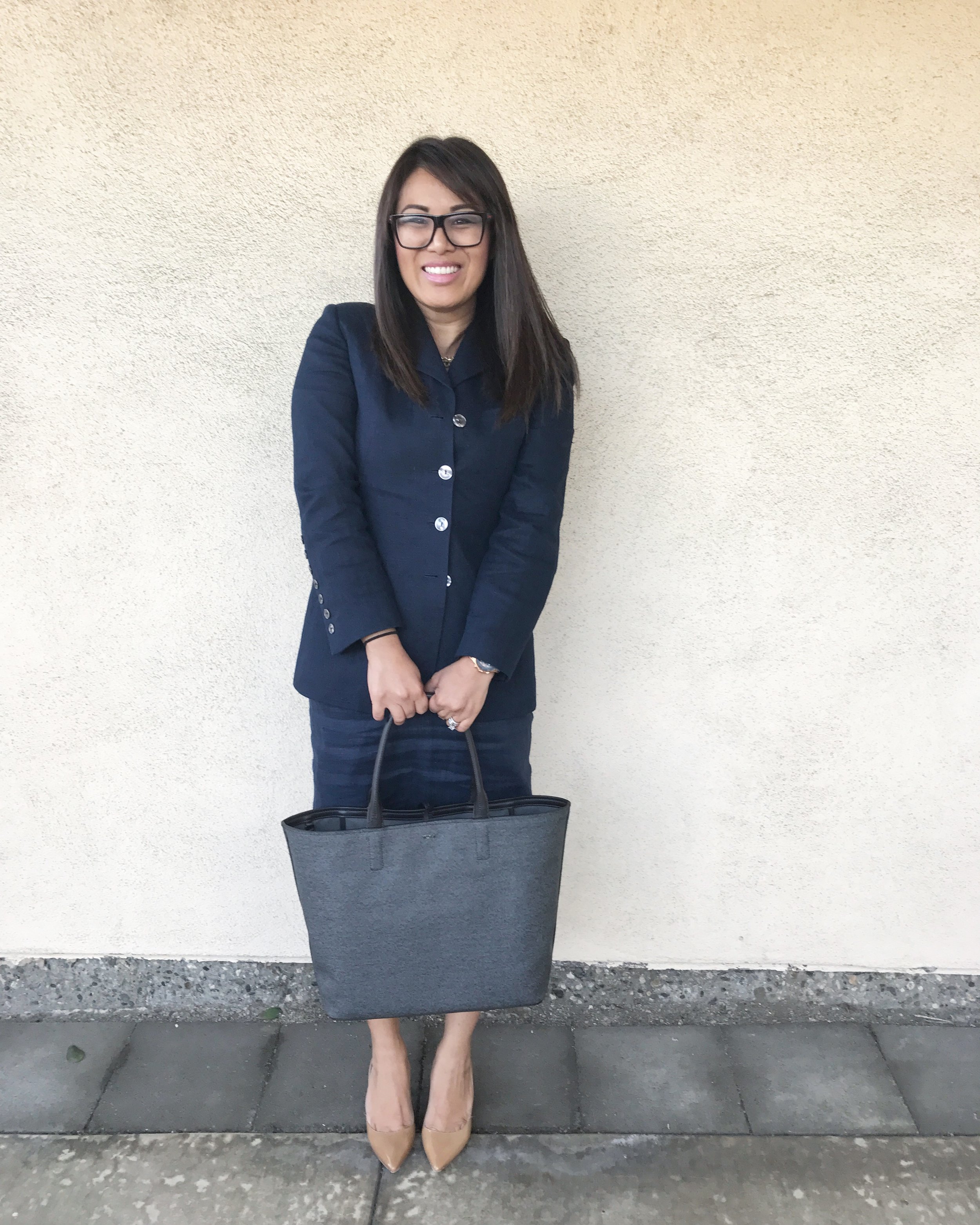 A woman with glasses and long dark hair, dressed in a navy blazer and skirt, holding a large gray tote bag and standing against a plain beige wall.