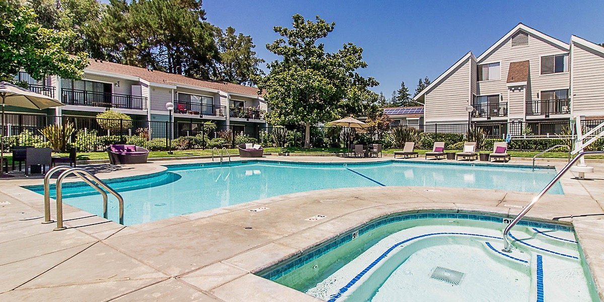 An outdoor pool area in an apartment complex, with lounge chairs, umbrellas, and a hot tub, surrounded by trees and residential buildings.