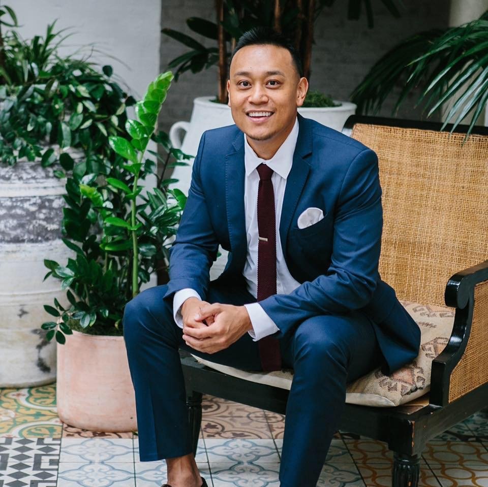 A man in a navy blue suit, white shirt, and maroon tie sitting on a bench in a setting with green potted plants and patterned tile flooring.