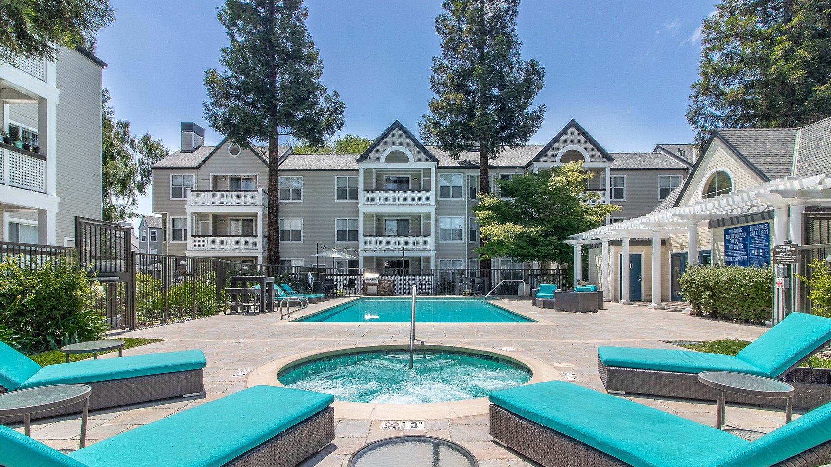 Outdoor swimming pool with lounge chairs, surrounded by apartment buildings, and a small hot tub in the foreground, under a clear blue sky.