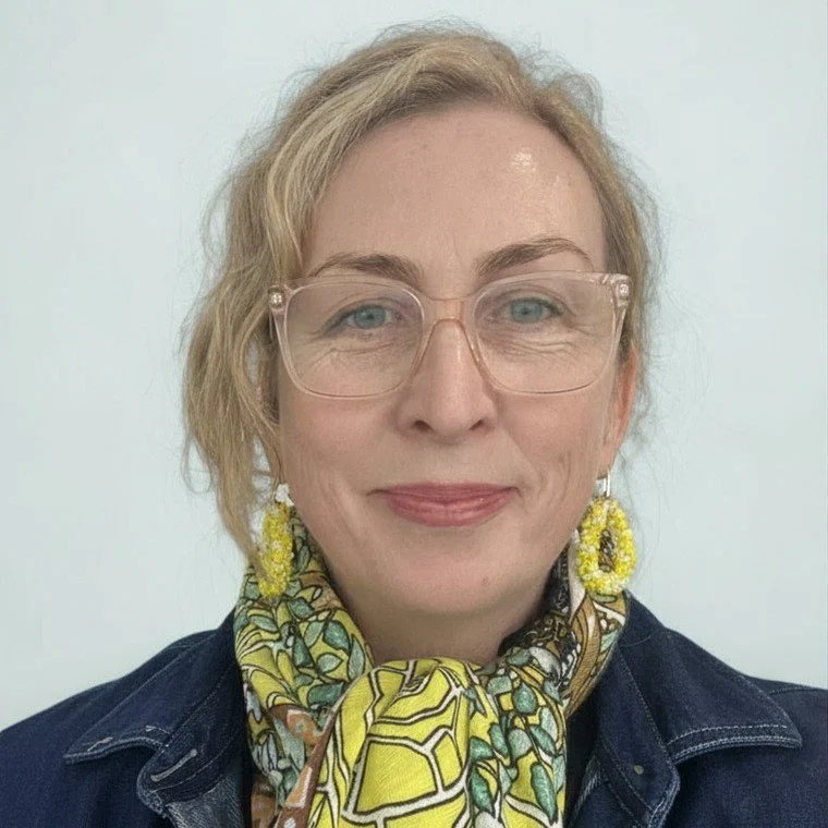 A woman with blonde hair, wearing glasses, yellow earrings, and a colorful scarf, smiles at the camera.