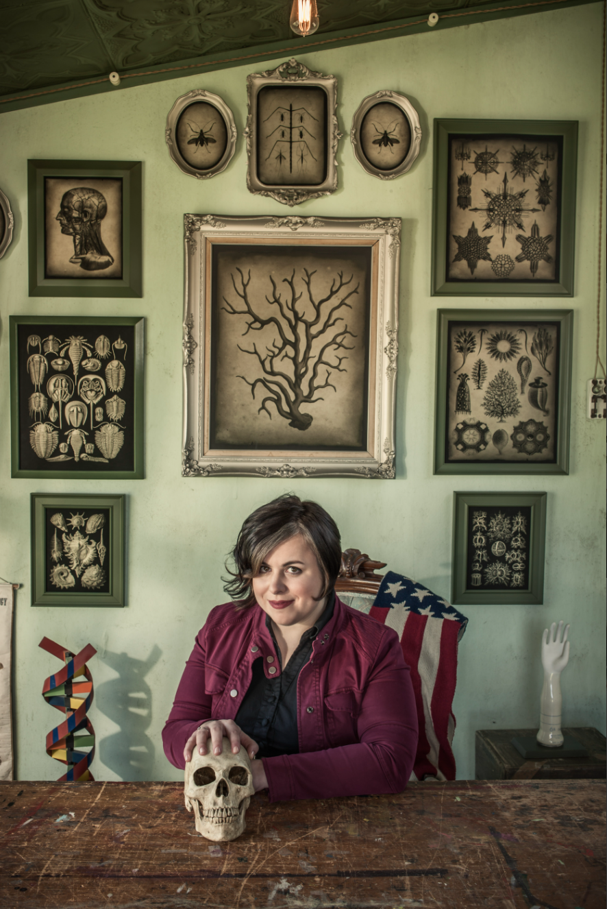 A woman with short dark hair and a maroon jacket sits at a wooden table, holding a skull, in front of a wall decorated with framed botanical and anatomical illustrations. An American flag blanket and a mannequin hand are also visible in the background.