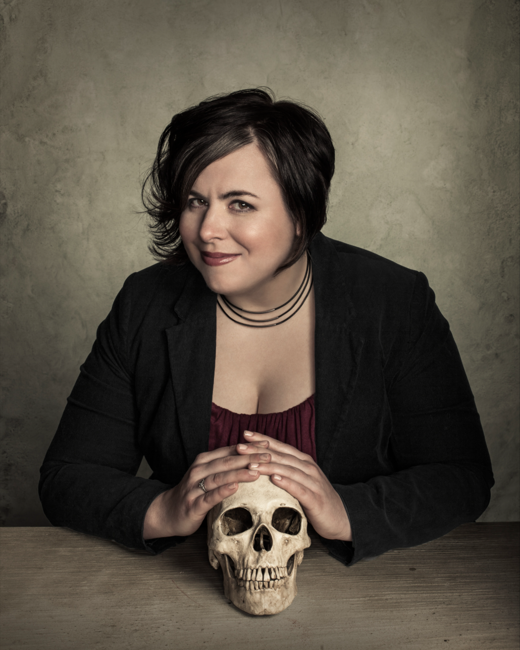 A woman with short dark hair and a smirk sits at a wooden table, resting her hands on a human skull.
