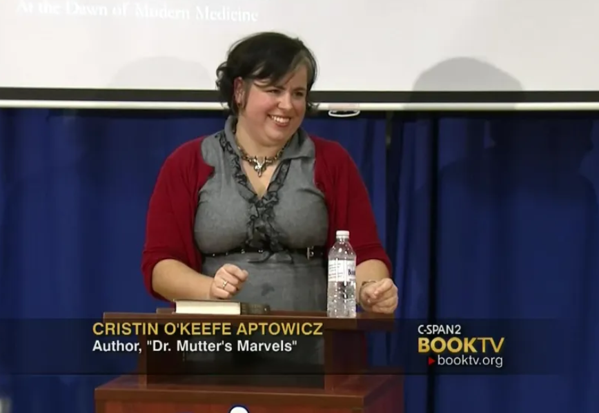 A woman with dark hair, wearing a gray dress and a red cardigan, is sitting at a table. She is smiling, with a bottle of water and a nameplate in front of her. Behind her is a blue curtain and a projection screen. The screen includes text about her, indicating her name as Cristin O'Keefe Aptowicz and her book 'Dr. Mutter's Marvels,' on C-SPAN2 Book TV.