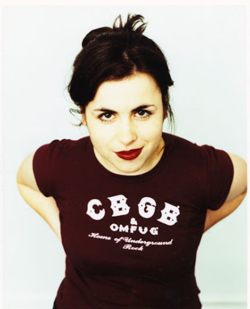 A woman with dark hair styled in a messy updo, wearing red lipstick, and a burgundy T-shirt with white text and graphics that say 'C.B.B OMFUG' and 'House of Underground Rock'.