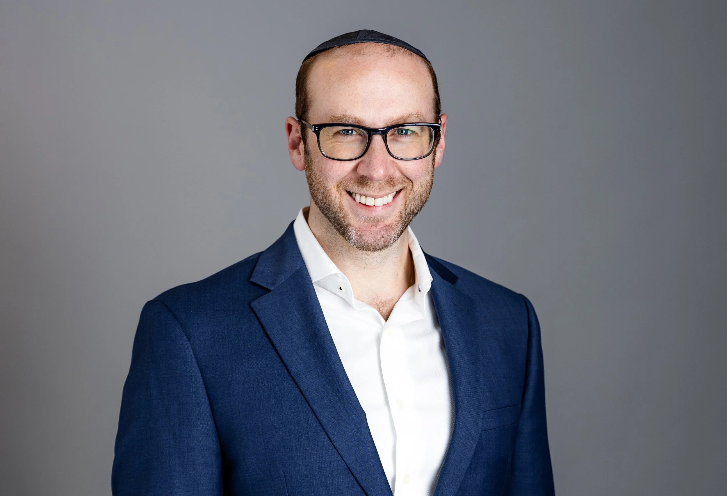 A smiling man wearing glasses, a dark blue suit, and a white shirt, standing against a gray background.