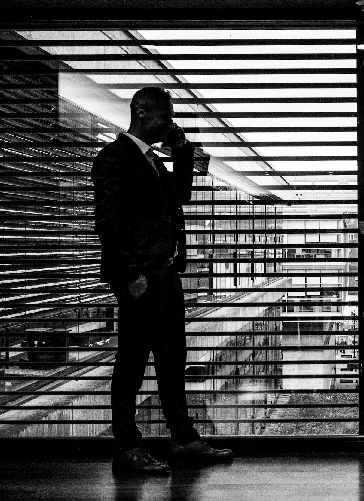 Silhouette of a man in a suit talking on a cellphone, standing in front of a window with horizontal blinds, inside a modern building.