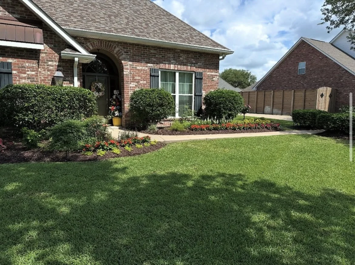 Front yard with green grass, landscaped flower beds, and bushes in front of a brick house with a window and a decorated porch, under a partly cloudy sky.