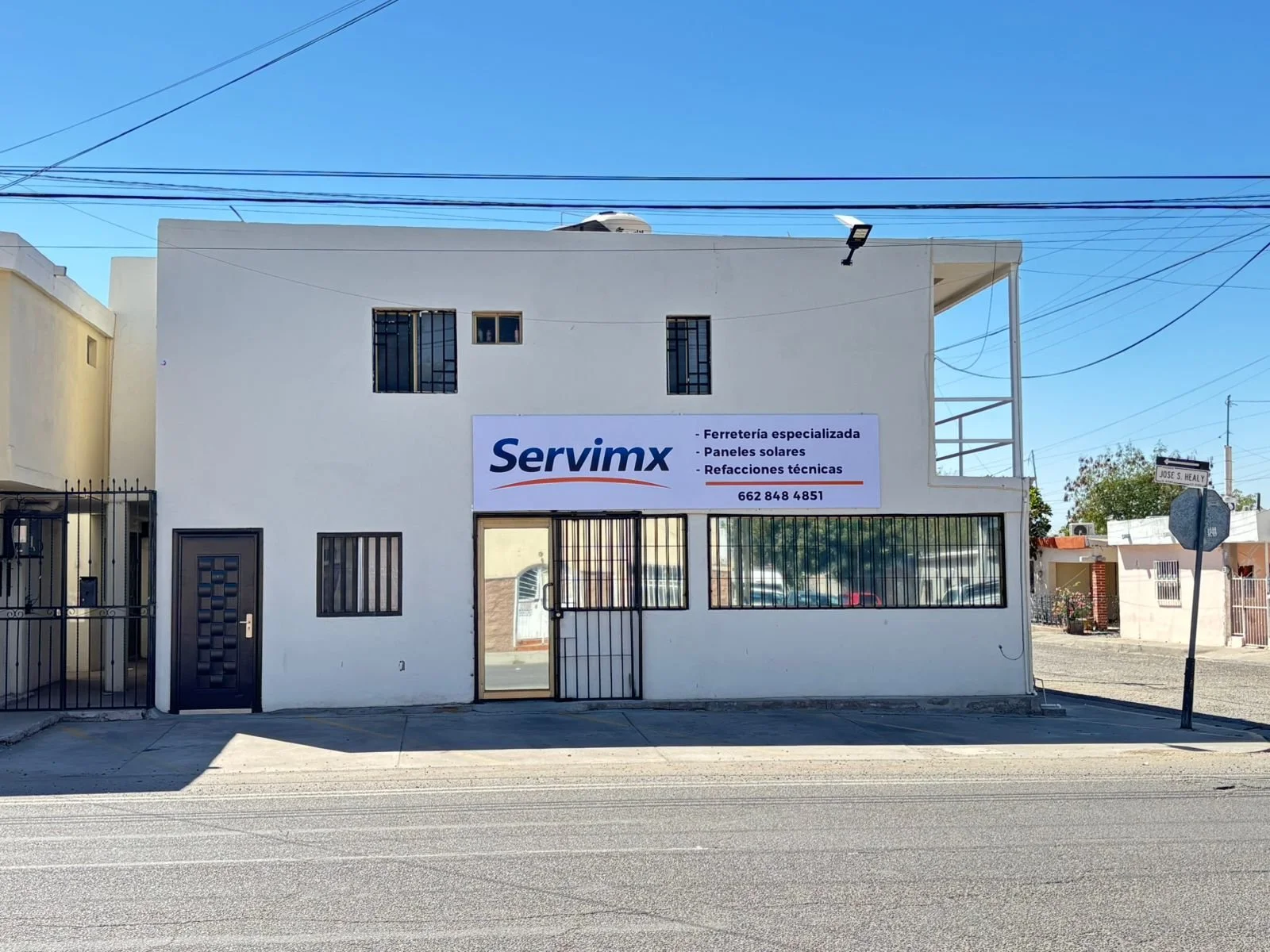 Edificio blanco con un cartel que dice Servimx, ofrecen ferretería especializada, paneles solares y refacciones técnicas, con ventanas y reja en la puerta principal, en una calle con líneas en la carretera y un poste con una señal de tránsito.