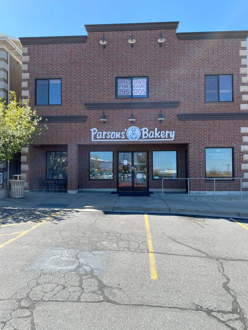 Front of a brick building with a bakery sign named Parsons Bakery, with an office space for lease sign in a second story window, parking lot with yellow lines and cracked pavement, a small outdoor seating area with chairs and a trash can, some trees,