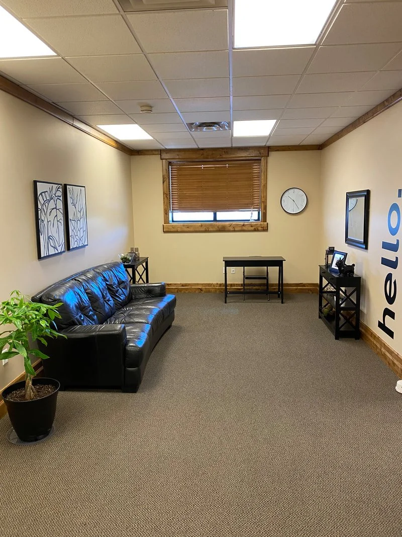 An office waiting room with a black leather couch, potted plant, framed artwork, a small table, a wall clock, a window with blinds, and a wall with large blue text reading "hello".