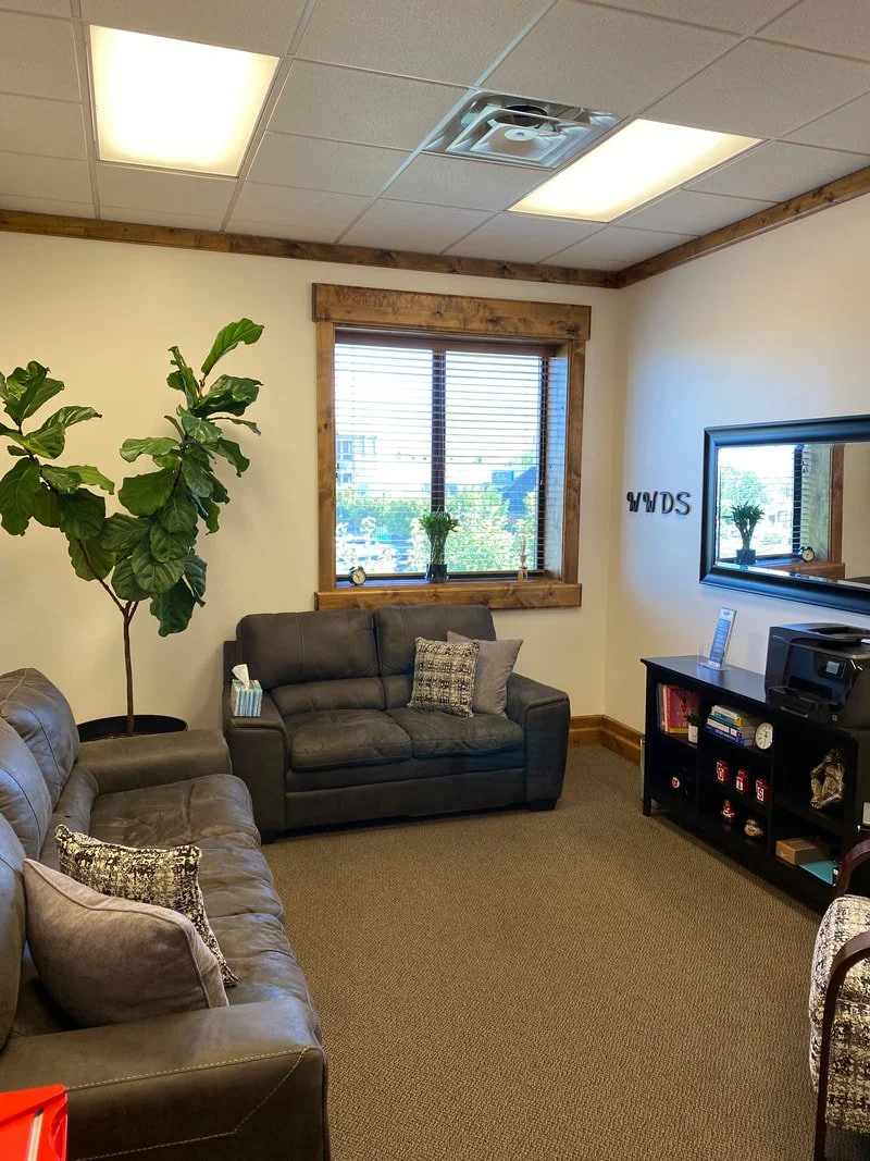 A waiting room with two dark gray sofas, a large green plant, a window with blinds, a small black shelving unit, and a wall mirror. The room has beige walls, carpet, and wooden trim.