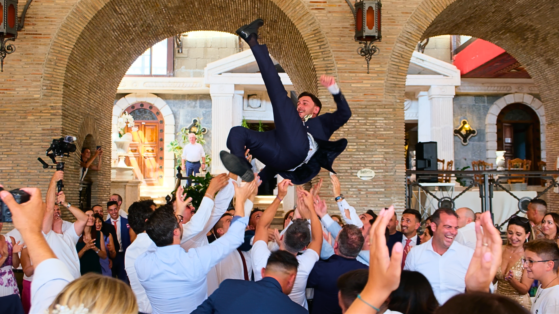 Invitati che sollevano lo sposo durante i festeggiamenti in una location storica con archi in mattoni. Animazione coinvolgente e party di matrimonio MDM Eventi