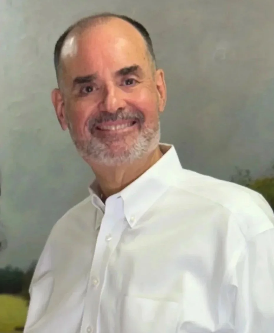 Dr. Benny Fernandez, a middle-aged man with a beard and mustache, wearing a white button-up shirt, standing against a plain background.