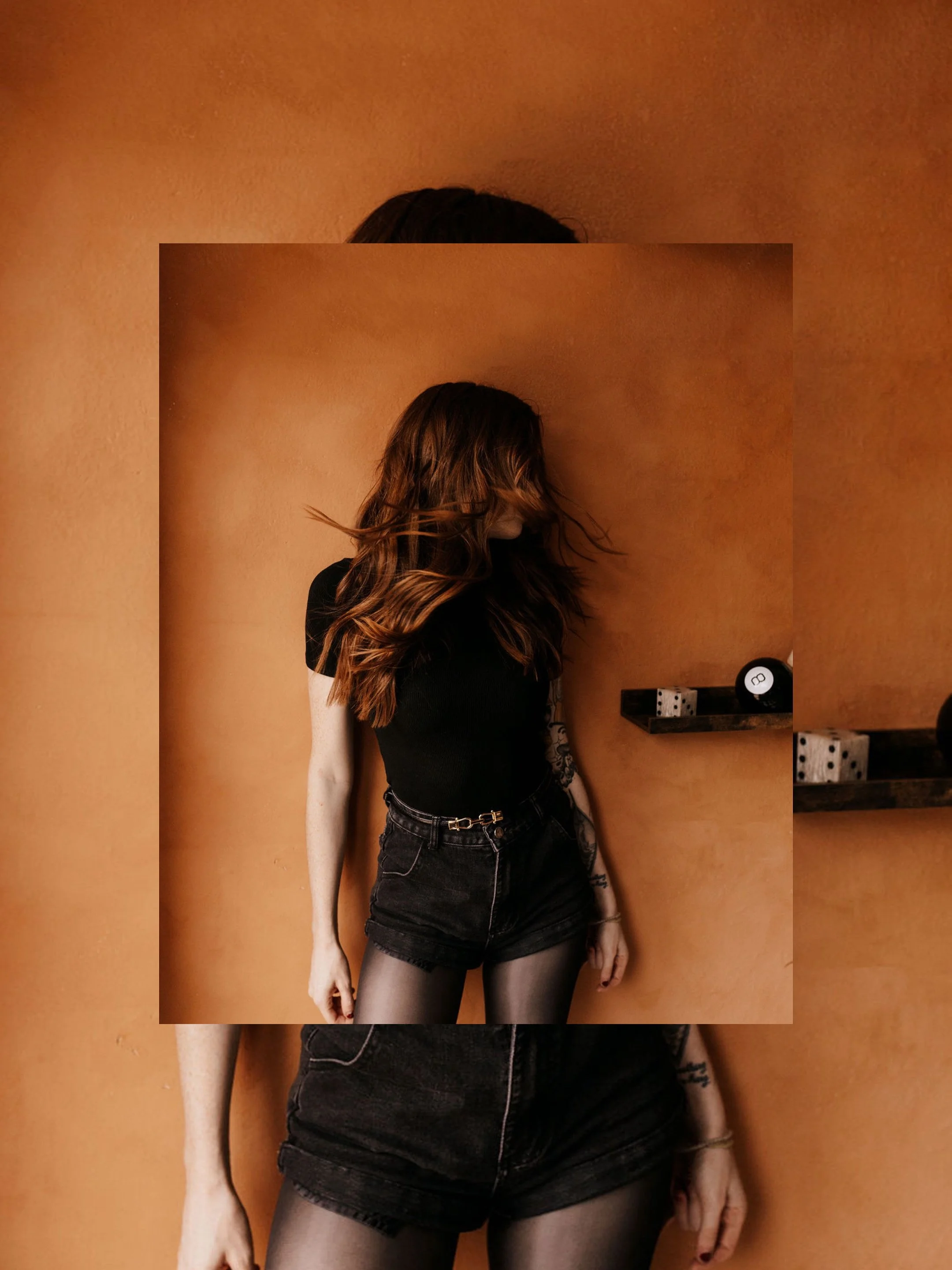 A woman with long wavy red hair, wearing a black t-shirt, denim shorts, and sheer black tights, standing against an orange wall with a shelf holding a dice and other objects.