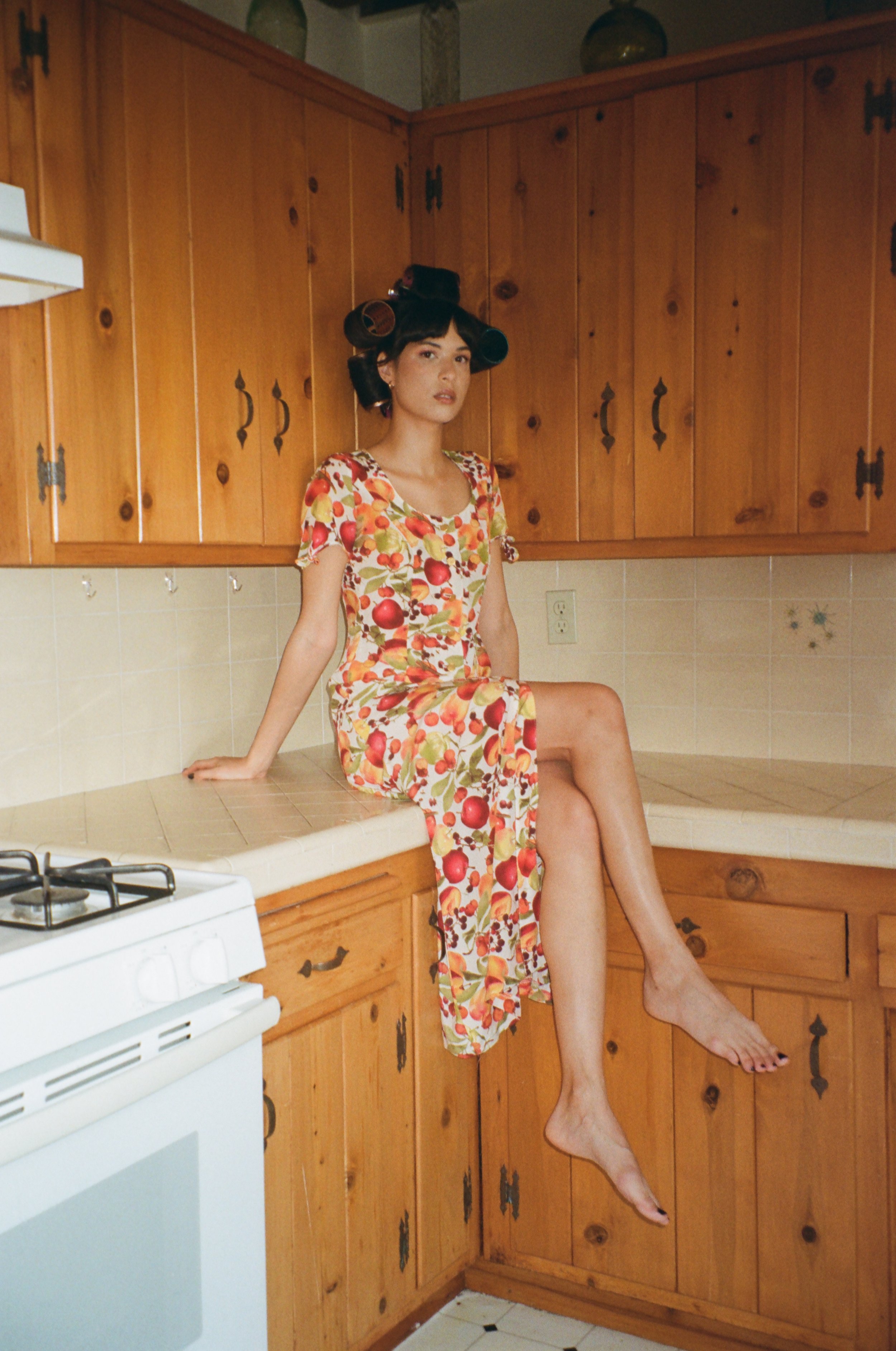 A woman in a colorful floral dress sitting on a kitchen counter with wooden cabinets and a stove in the background.