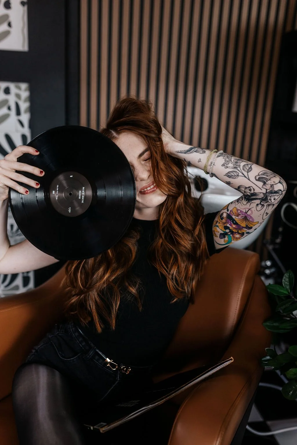A woman with wavy red hair and tattoos on her arms holding a black vinyl record in front of her face, sitting on a brown leather couch in a modern room.