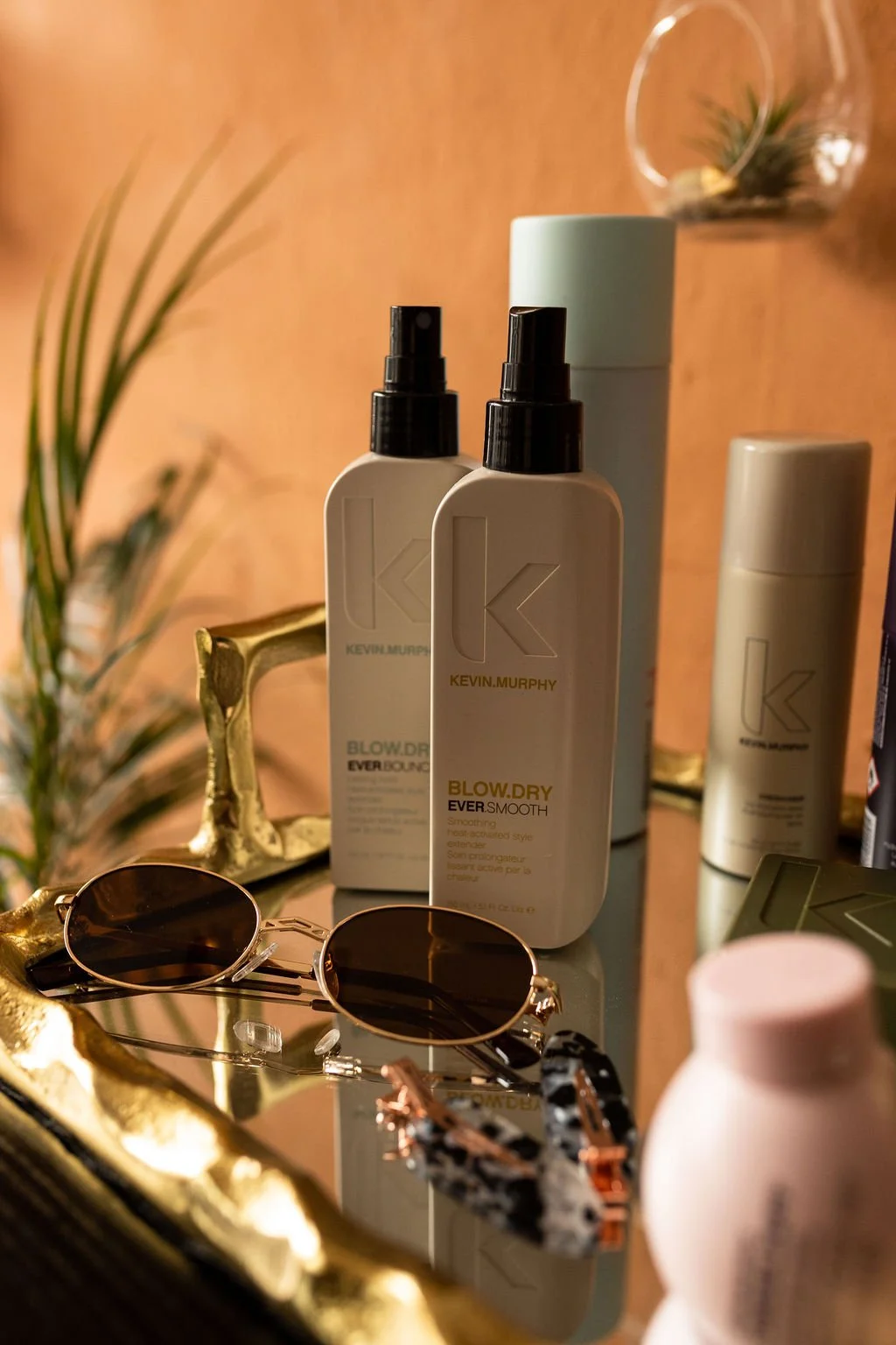 Kevin Murphy Blow Dry spray bottles with sunglasses, gold decoration, and a plant in the background.