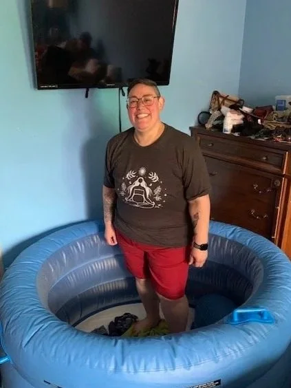 Person standing inside a blue inflatable pool, smiling, in a room with a wooden dresser and a wall-mounted TV.