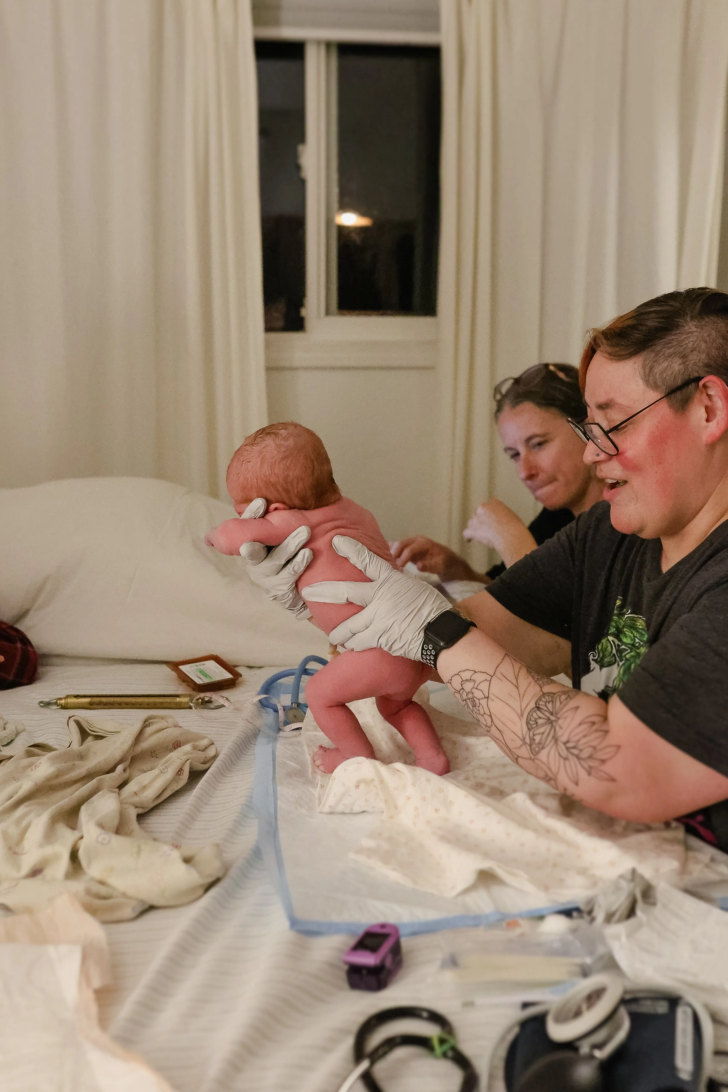 A person holding a newborn baby during childbirth in a hospital room. Two women are sitting in the background, one smiling, with medical equipment and supplies on the bed.