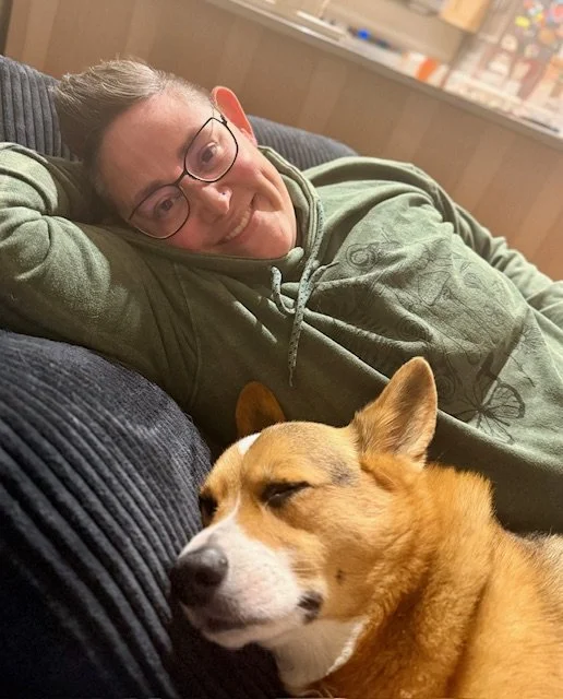 A person with glasses lying on a couch with a green hoodie, smiling, and a sleeping dog with brown fur resting nearby.