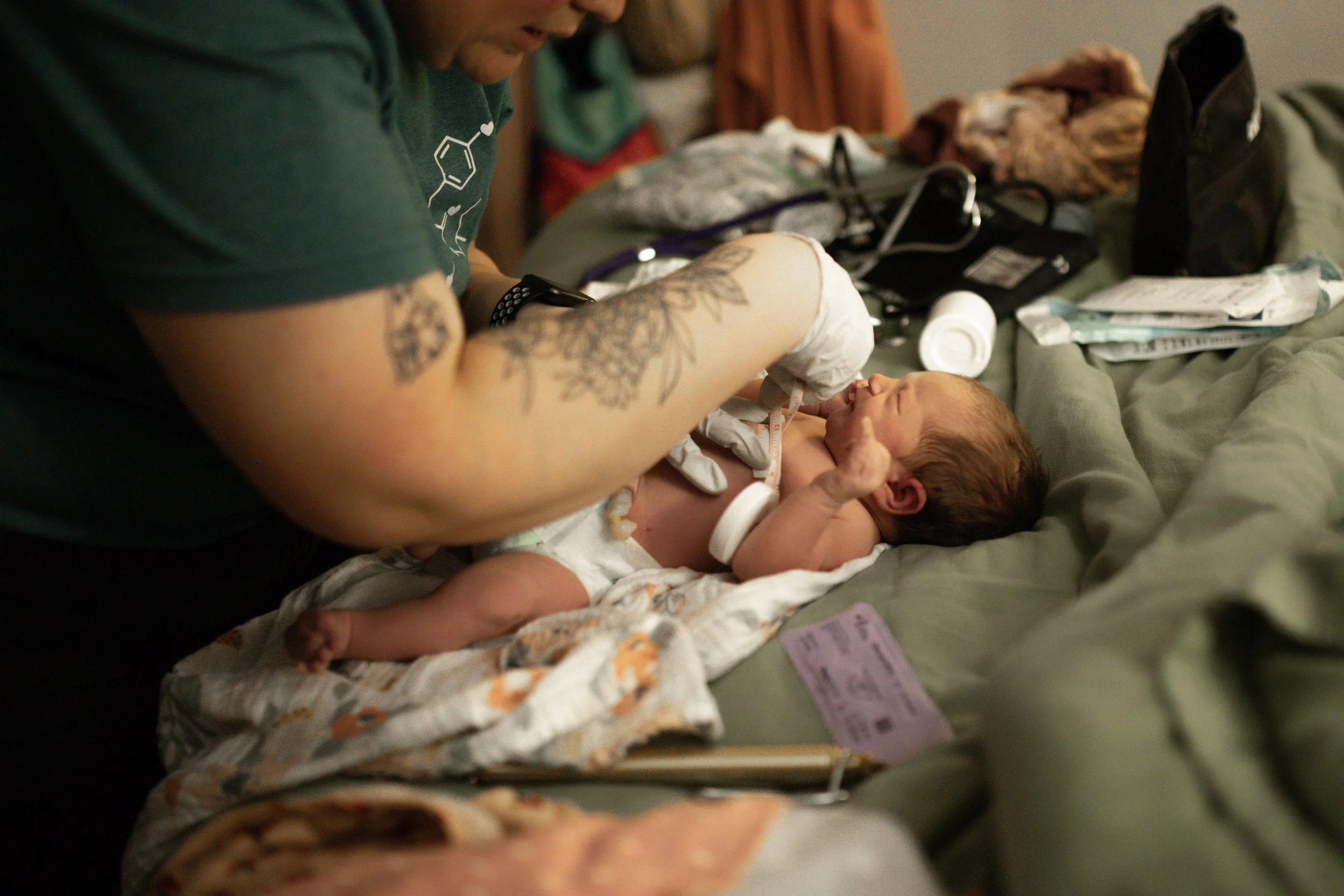 A person with tattoos is feeding a newborn baby with a bottle in a hospital or medical setting, with medical supplies and a hospital sheet around.