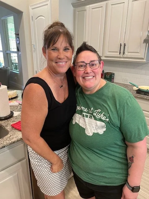 Two women smiling in a kitchen. One woman has brown hair in a bun, wearing a black sleeveless top and patterned shorts. The other woman has short hairstyle, glasses, and tattoos on her arm, wearing a green T-shirt and black shorts.