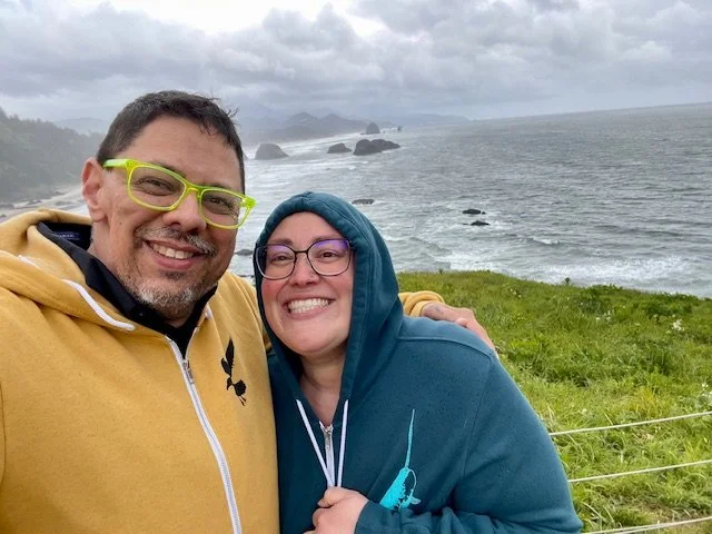 Two people smiling and embracing near a coastal landscape with grassy foreground and rocky shoreline in the background, overcast sky.