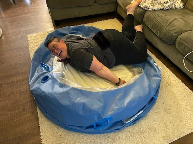 Person lying on their stomach on an inflatable pool float inside a living room, smiling, with a couch and a wooden floor nearby.