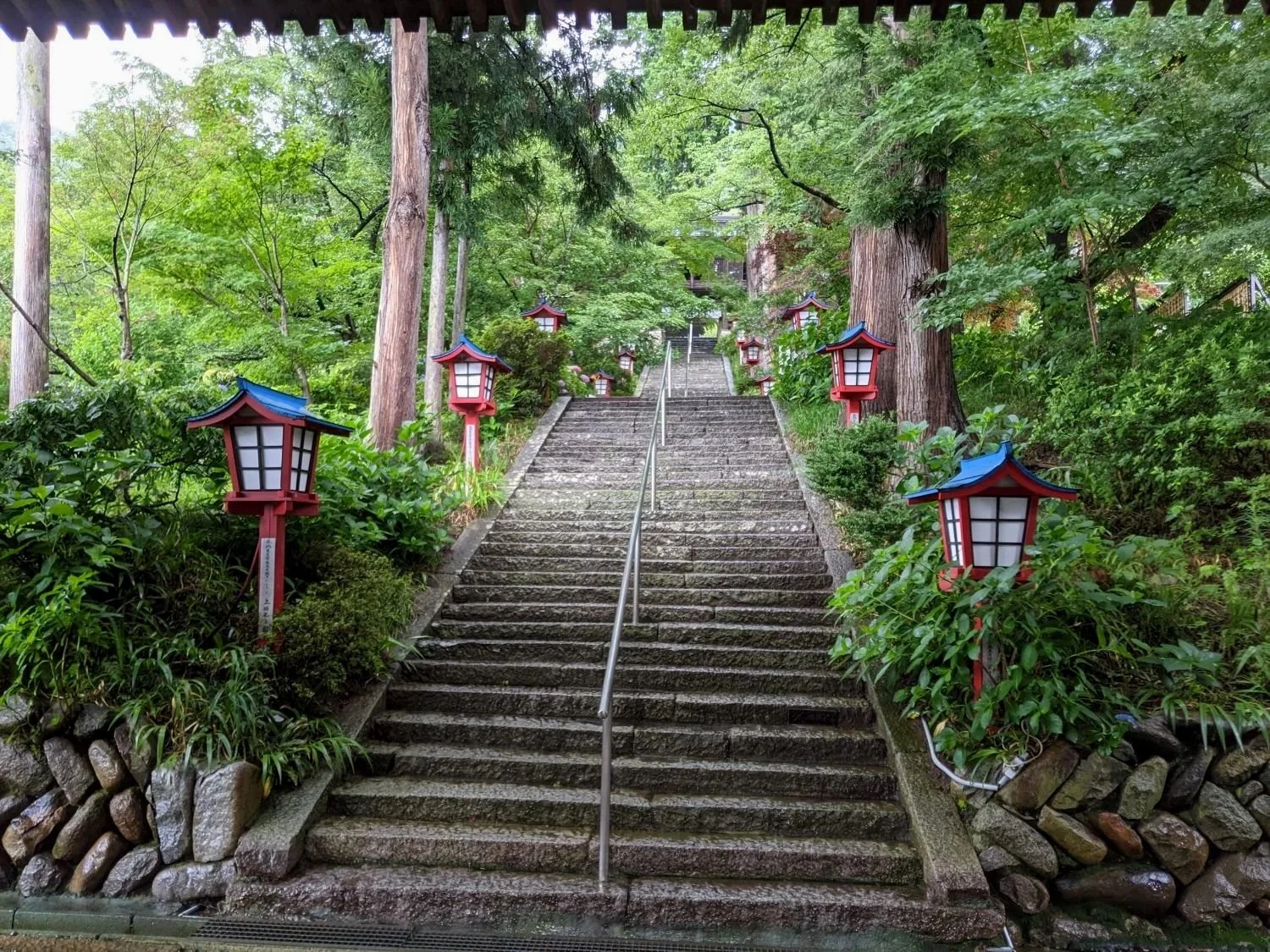Stone staircase of an ancient temple in Yamanashi Japan, with red and blue Japanese-style lanterns on either side, surrounded by green trees and shrubs.
