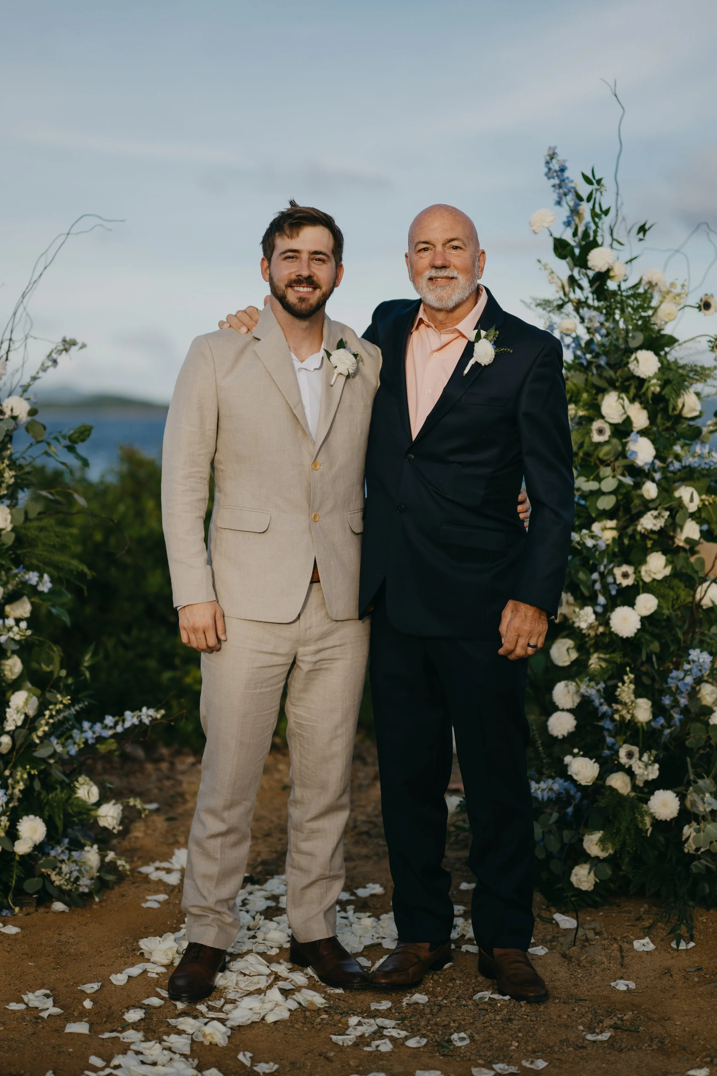 Two men (father and son) in suits standing outdoors near a floral arch, smiling at the camera, with ocean and sky in the background.
