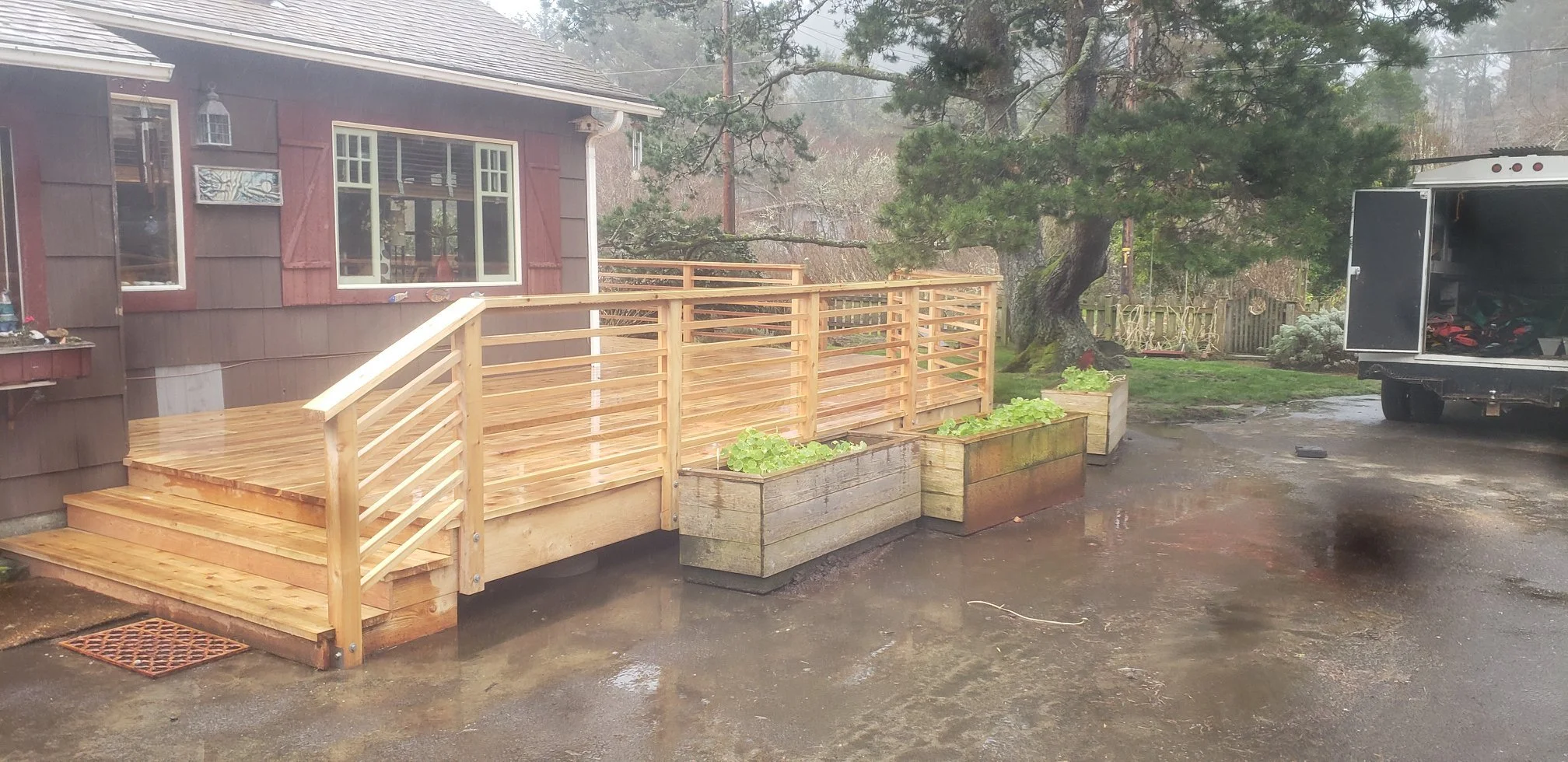A newly built wooden deck attached to a house on the Oregon Coast, with steps leading up to it. In front of the deck, there are three rectangular wooden planters with green plants.