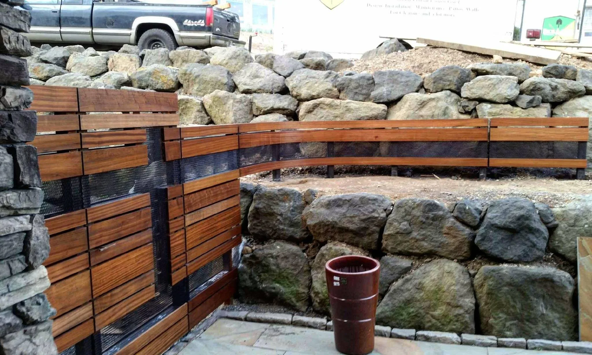 A garden with a stone wall and a wooden slat fence on the Oregon Coast, with a red flowerpot on the ground and a truck in the background.