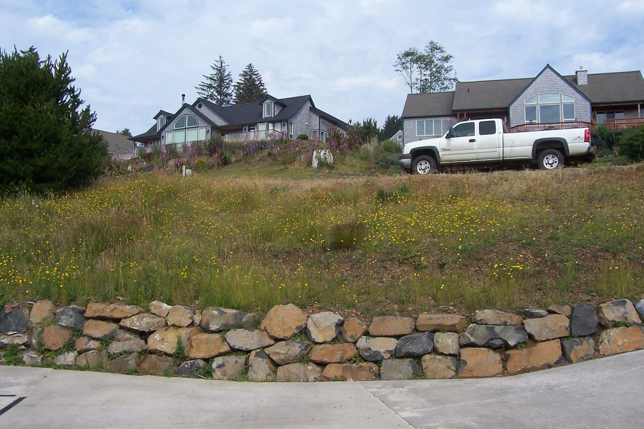 A residential area with two houses on a hill on the Oregon Coast, a white pickup truck parked on the driveway, and a flower garden with yellow and purple flowers growing along a stone border. The foreground has a concrete sidewalk.