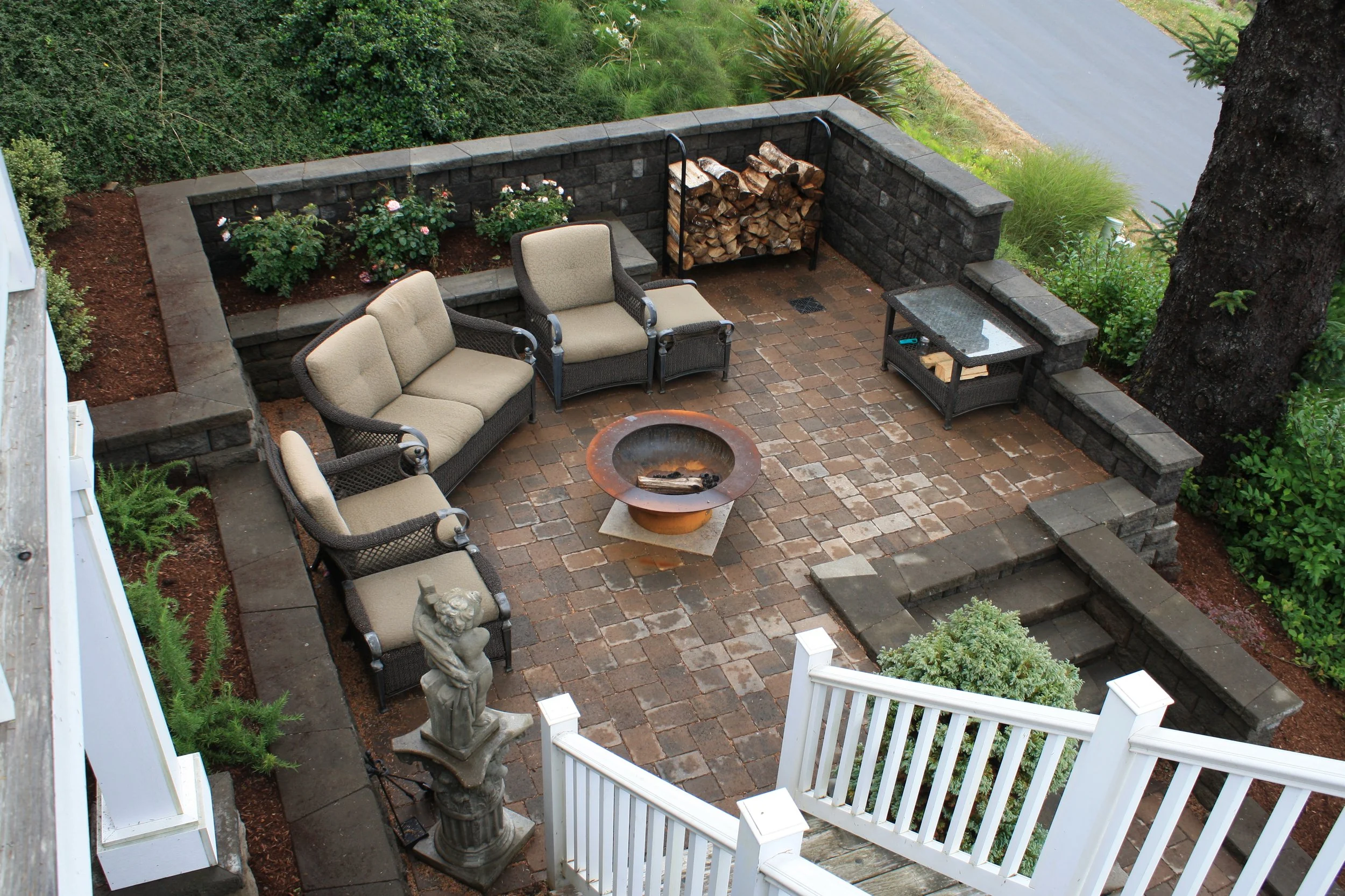 A backyard patio on the Oregon Coast with brick flooring, patio furniture, a fire pit, a stone firewood rack, and a garden border with blooming flowers, surrounded by lush greenery and trees.