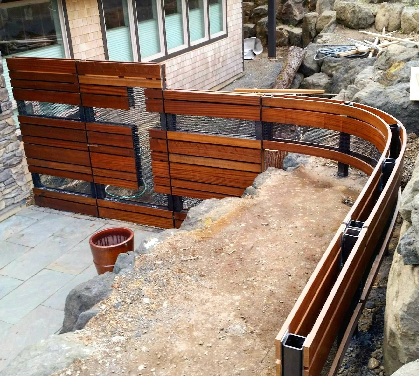 A wooden and metal fence under construction on the Oregon Coast next to a house with stone exterior walls. The fence has wooden slats with black metal mesh panels and is curved.