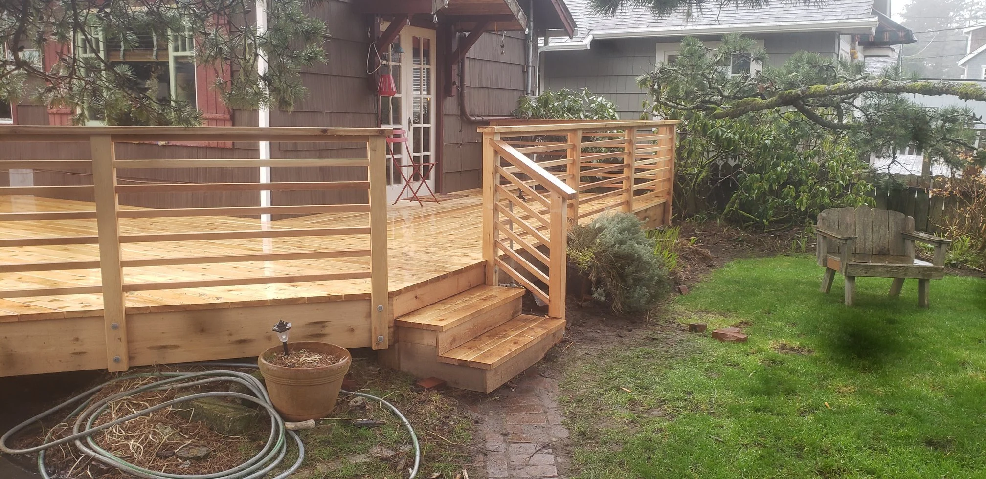 A newly built wooden deck attached to the back of a house on the Oregon Coast, with steps leading down to a grassy yard. In the yard, there's a weathered wooden bench and some garden hoses on the ground.