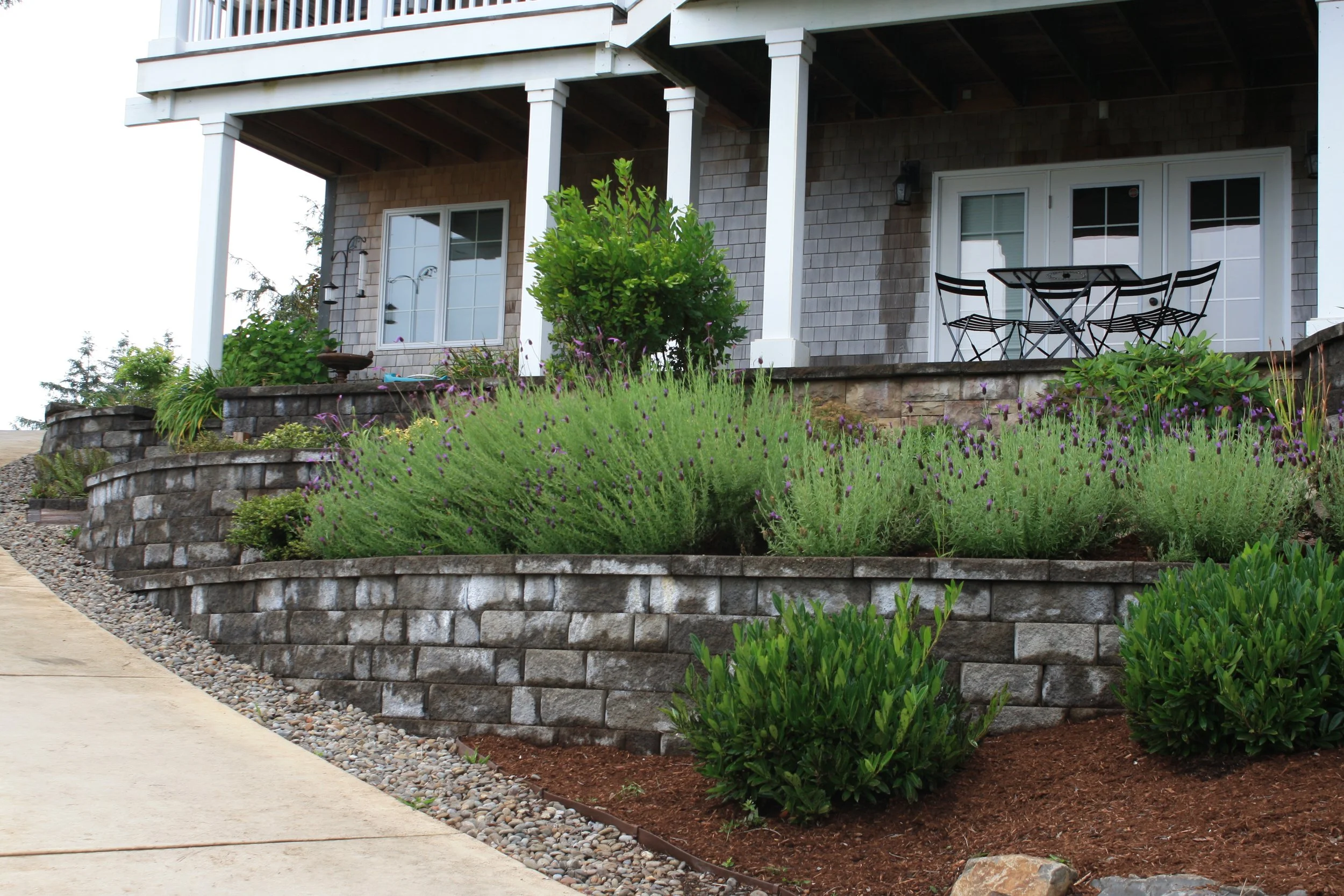 Front porch of a house on the Oregon Coast with steps and a small outdoor table with chairs, surrounded by a landscaped garden with various plants and flowers.