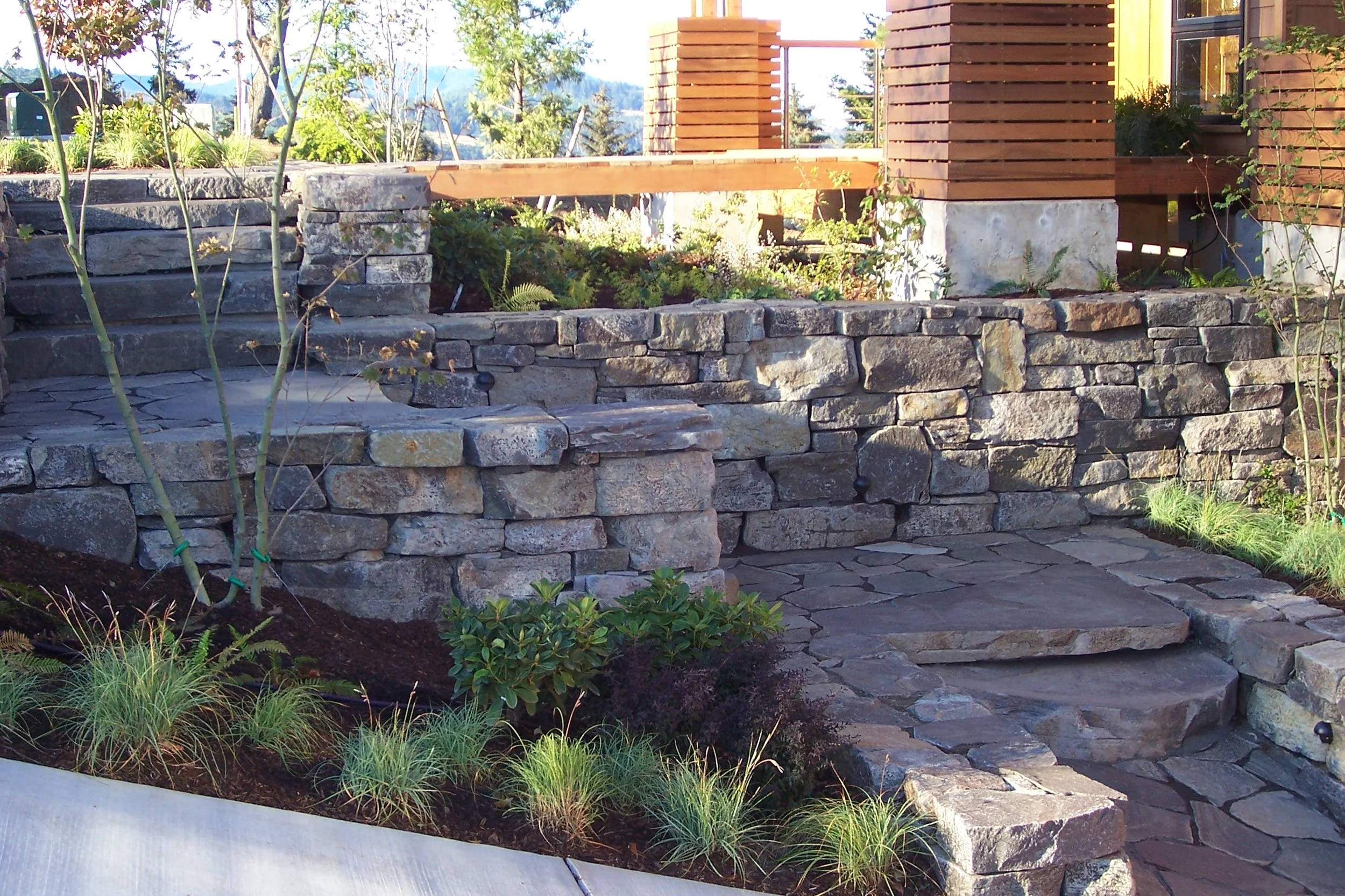 Side view of a backyard garden with a stone retaining wall, built-in stone steps, a wooden deck, and various plants. Includes trees, shrubs, and grass with scenic mountains in the background.