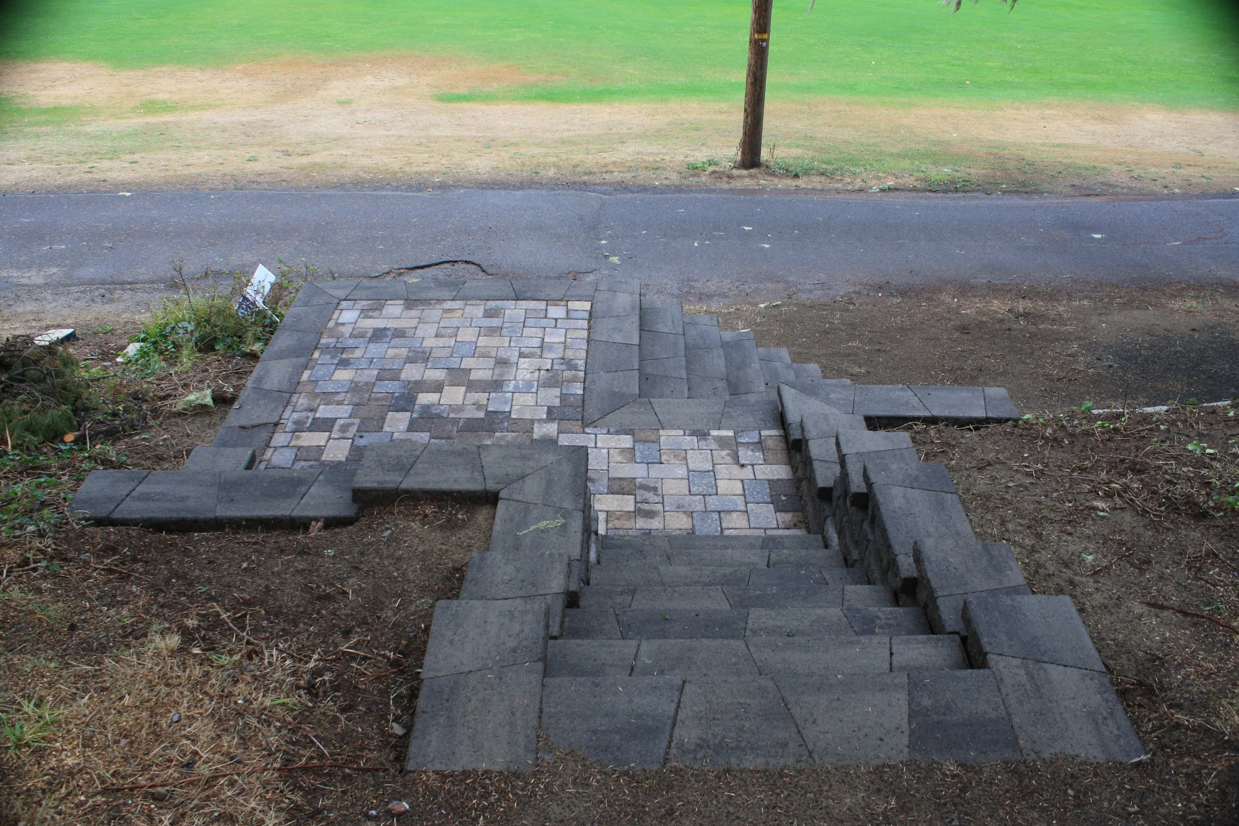 A set of outdoor stairs on the Oregon Coast made of gray bricks leading to a sidewalk with a grassy area and a tree in the background.