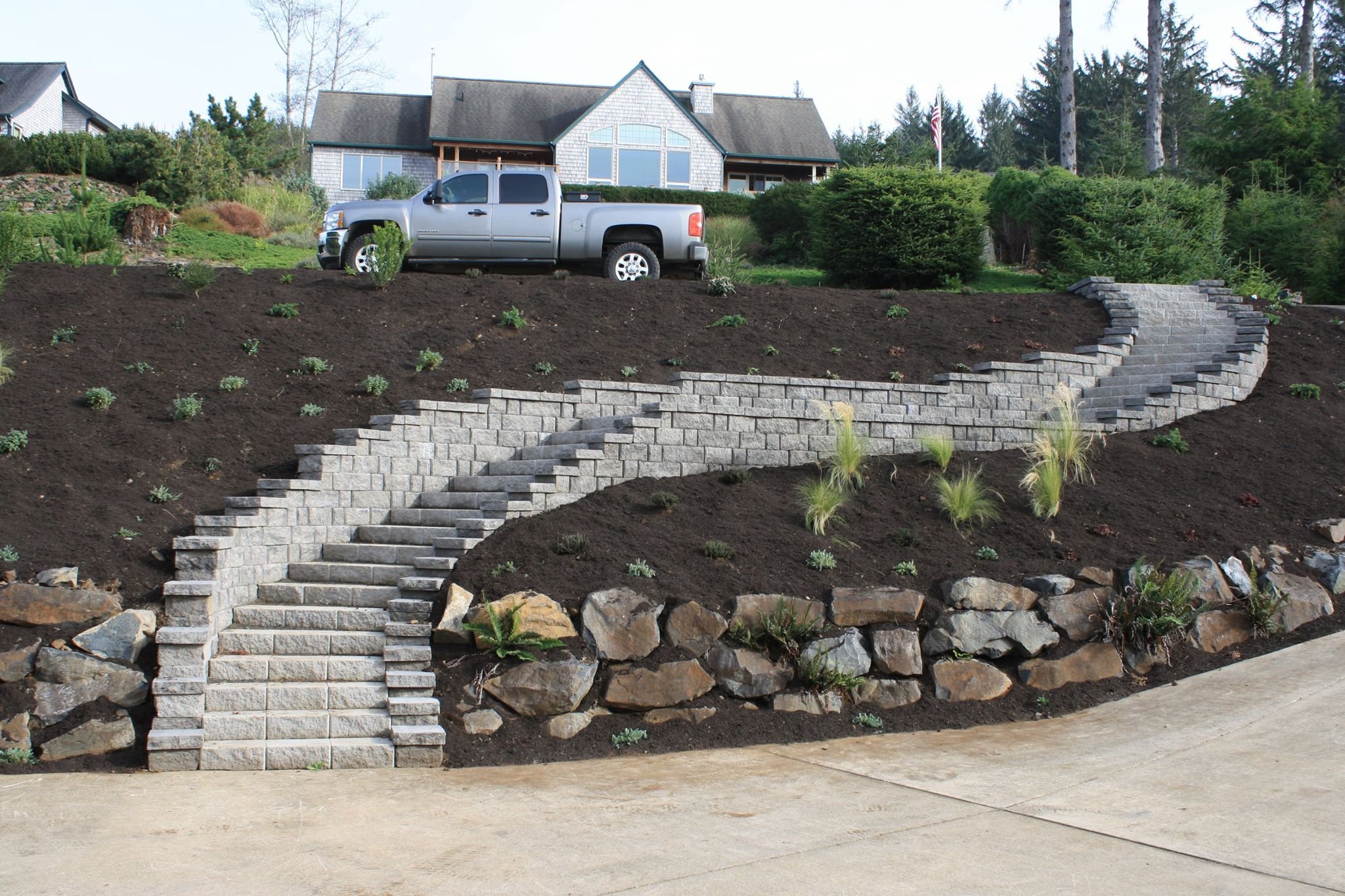 A house on a hill with a landscaped garden on the Oregon Coast, a gray truck parked in front, and a staircase made of stone blocks leading up to the house.