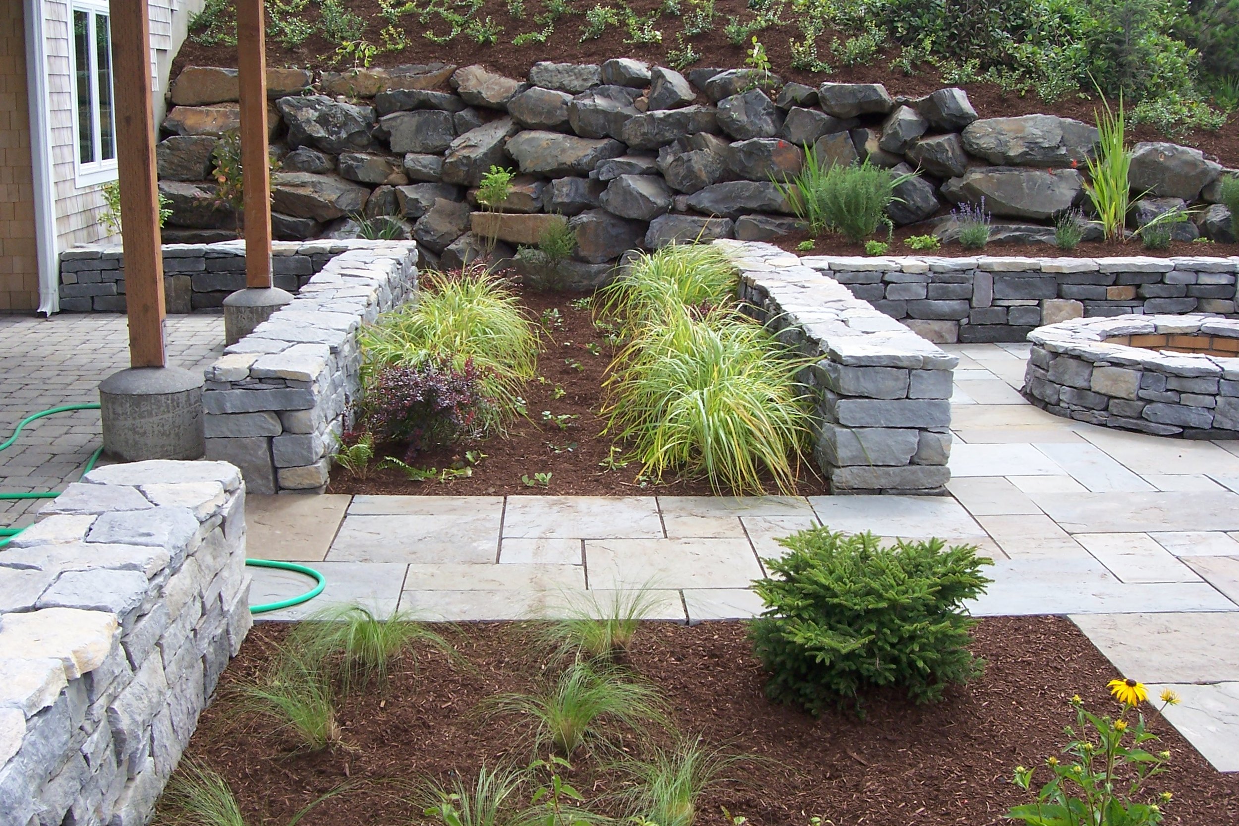 A landscaped garden on the Oregon Coast with tiered stone retaining walls, various plants, and a paved patio area.