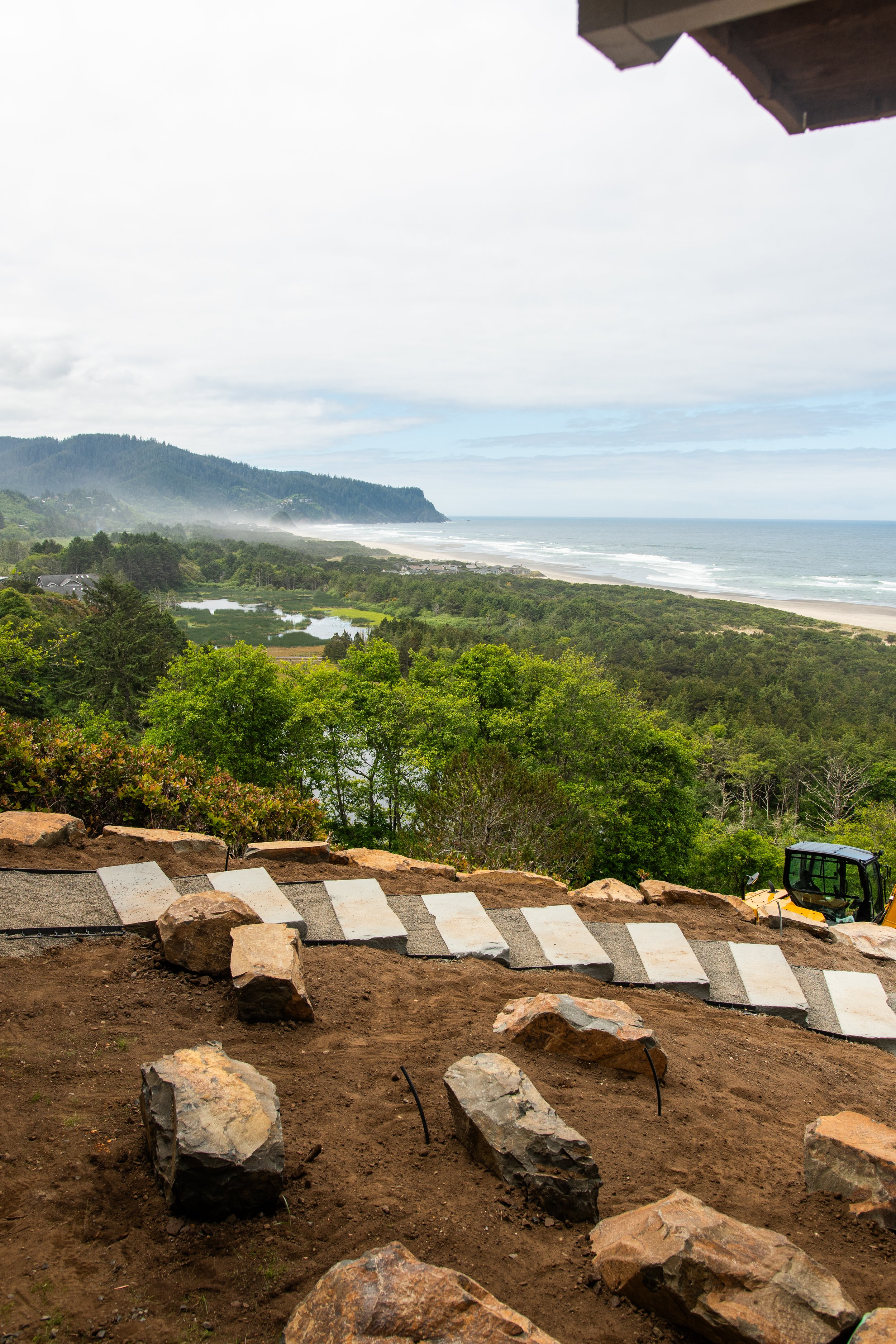 A construction site on the Oregon Coast with rocks and dirt, in the foreground, overlooking a scenic view of a grassy landscape, ocean, and hills under a cloudy sky, with part of a building and construction equipment visible.