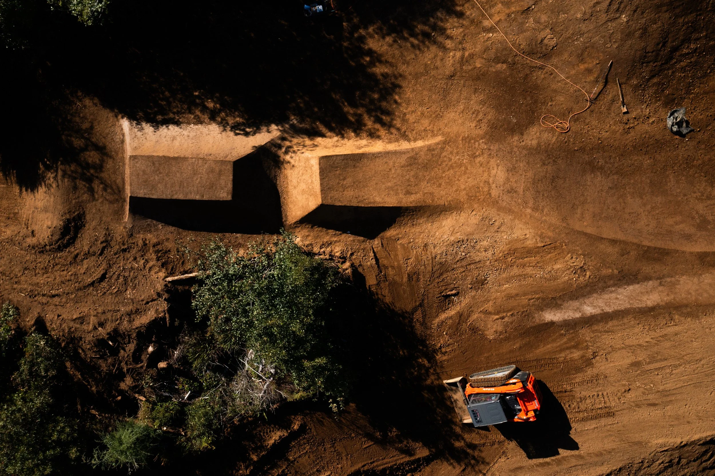 Nighttime scene of an excavation site with a large orange excavator and a partially dug earth mound. There are green bushes and trees at the edge of the site, and construction tools are visible on the ground.